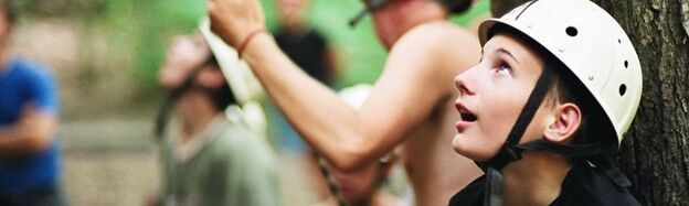 The image shows a young person wearing a white helmet, looking upwards with an expression of surprise or awe. Other people are visible in the background, suggesting an outdoor activity or adventure. The setting appears to be a forest or natural environment.